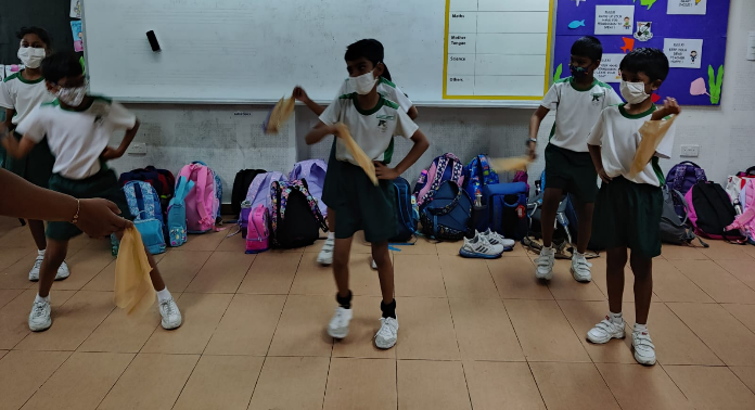 Four masked school children in white and green uniforms dance with scarves in a classroom with backpacks along the wall.